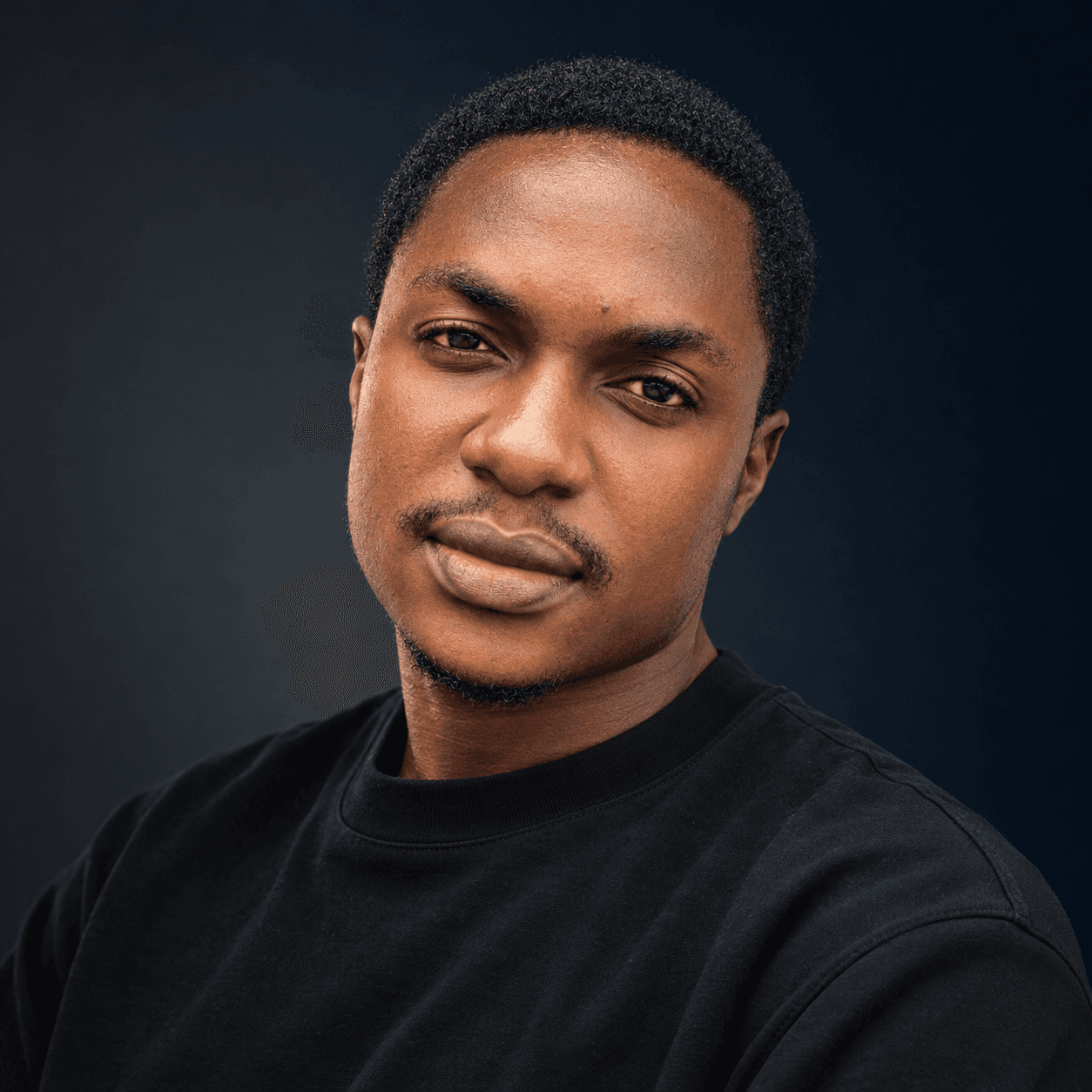 Portrait of a Black man with short hair, wearing a black t-shirt against a dark background.